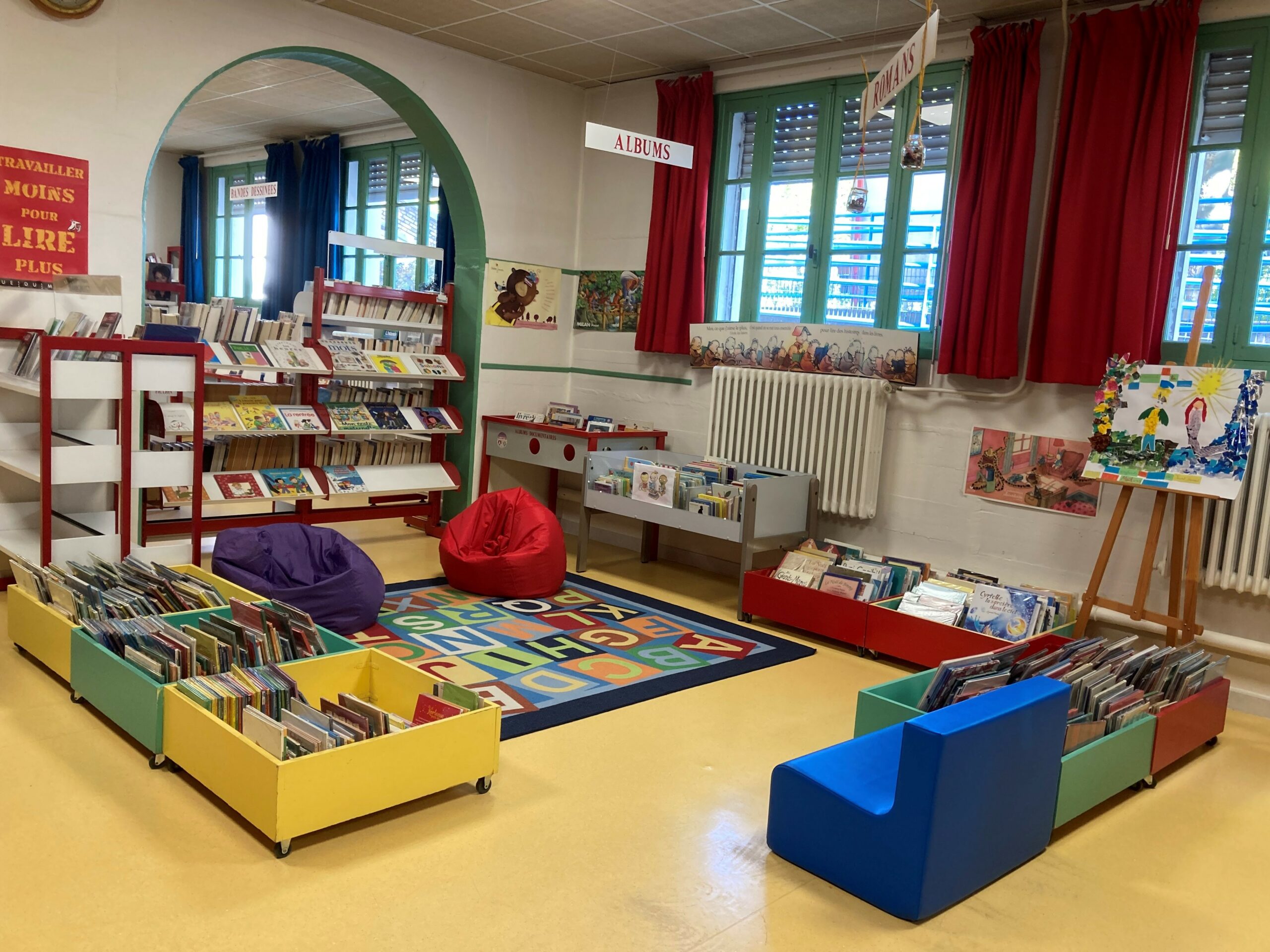 Médiathèque Photo de l'intérieur de la médiathèque avec un tapis carré au sol représentant les lettres de l'alphabet de façon coloré, entouré par des bacs à livres et des poufs pour s'assoir.