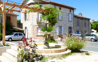 Photo de la place Augustin Malroux avec sa fontaine, une pergola végétalisée et une façade de maison en pierre apparentes.